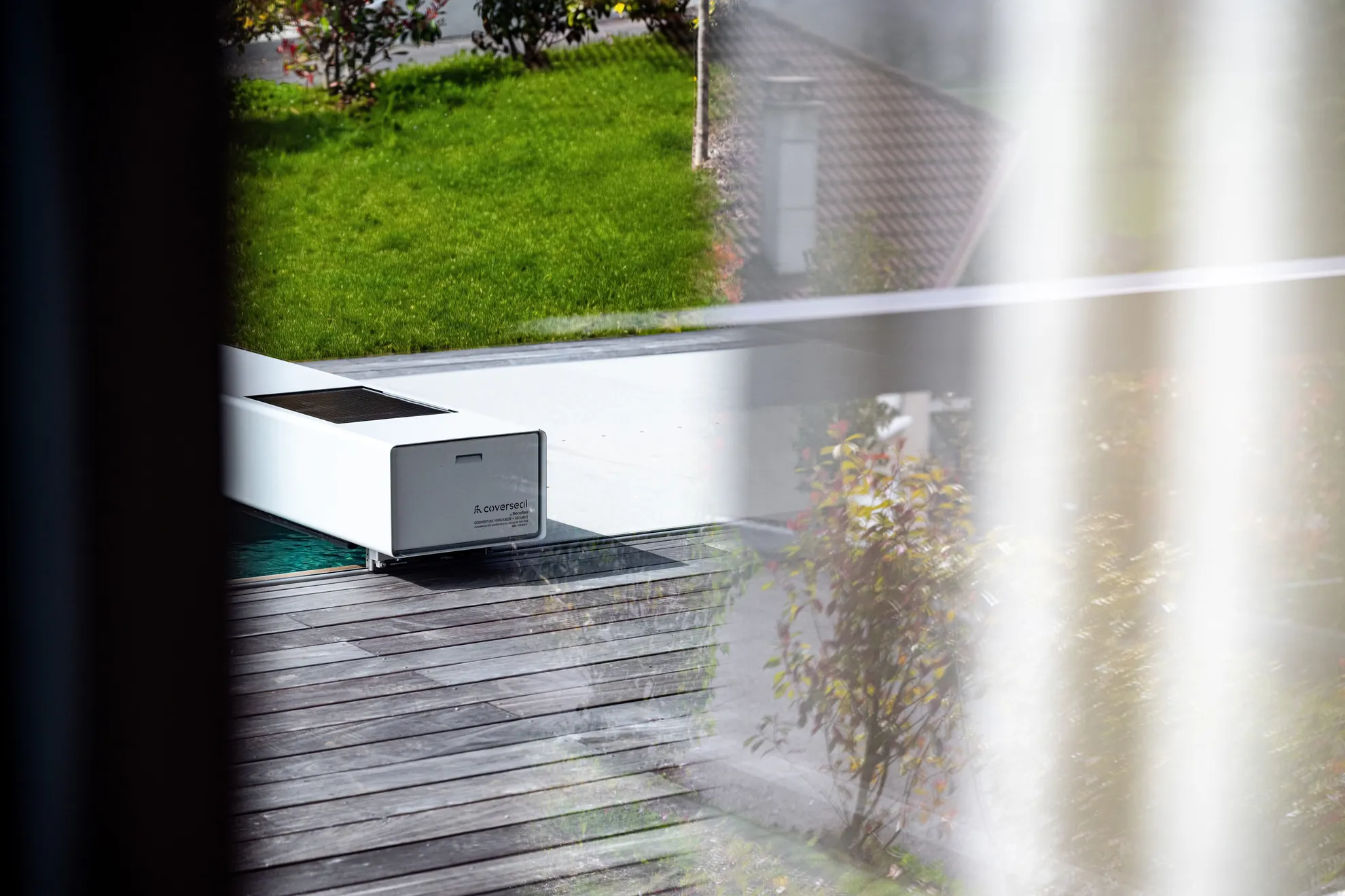 Couverture de piscine Coverseal grise avec habillage gris clair, intégrée à une terrasse en bois bordée de verdure.